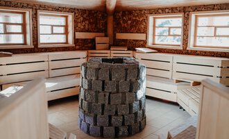 Ein gemütlicher Saunaraum mit Holzbänken und großen Fenstern. In der Mitte steht ein Ofen aus Steinen. | © Verwöhn- & Wellnesshotel Walserhof| Aileen Melucci