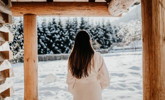 Eine Frau in einem weißen Bademantel steht unter einem Holzvordach und schaut auf eine verschneite Landschaft. Die Bäume im Hintergrund sind mit Schnee bedeckt. | © Verwöhn- & Wellnesshotel Walserhof| Aileen Melucci