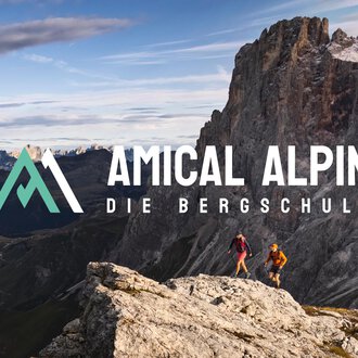 © SALEWA | Fotograf Storvteller Labs Zwei Personen wandern auf einem Felsen mit Blick auf beeindruckende Berge. Der Logo von "Amical Alpin" ist prominent im Bild platziert. | © SALEWA | Fotograf Storvteller Labs