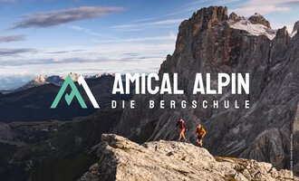 Zwei Personen wandern auf einem Felsen mit Blick auf beeindruckende Berge. Der Logo von "Amical Alpin" ist prominent im Bild platziert. | © SALEWA | Fotograf Storvteller Labs