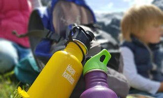 Two colorful water bottles are standing on green grass. In the background, children and mountains can be seen. | © Austrian Ski- & Service Ranch | Trixi Leitner