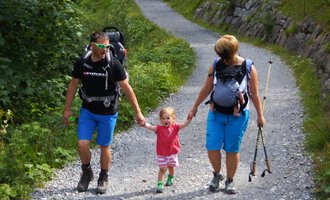 Eine Familie wandert auf einem schmalen Weg durch die Natur. Die Eltern halten die Hände ihres kleinen Kindes und tragen Rucksäcke. | © Austrian Ski- & Service Ranch | Trixi Leitner