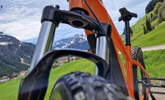 Ein orangefarbenes Mountainbike steht auf einem grünen Hügel mit Blick auf die Berge. Der Himmel ist klar und es ist ein sonniger Tag. | © Austrian Ski- & Service Ranch | Trixi Leitner