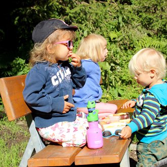 Three children are sitting on a bench outside. They are enjoying snacks and playing with toys on a table. | © Kleinwalsertal Tourismus | Britta Maier