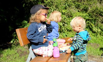 Three children are sitting on a bench outside. They are enjoying snacks and playing with toys on a table. | © Kleinwalsertal Tourismus | Britta Maier