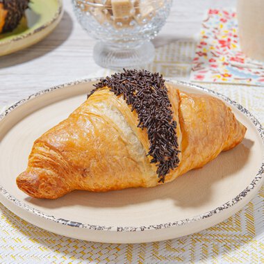 Ein köstlicher Croissant mit Schokoladenglasur liegt auf einem hellen Teller. Im Hintergrund sind weitere Frühstücksgegenstände sichtbar. | © MPREIS Warenvertriebs GmbH