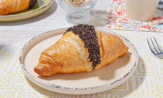 Ein köstlicher Croissant mit Schokoladenglasur liegt auf einem hellen Teller. Im Hintergrund sind weitere Frühstücksgegenstände sichtbar. | © MPREIS Warenvertriebs GmbH