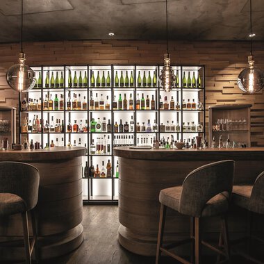Eine stilvolle Bar mit Holzinterieur und einem beleuchteten Regal voller Flaschen. Die Sitzgelegenheiten sind gemütlich und modern. | © Travel Charme Ifen Hotel
