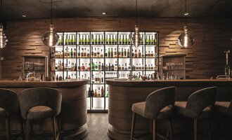 Eine stilvolle Bar mit Holzinterieur und einem beleuchteten Regal voller Flaschen. Die Sitzgelegenheiten sind gemütlich und modern. | © Travel Charme Ifen Hotel