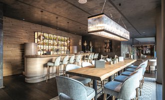 Ein eleganter Innenraum eines modernen Restaurants mit einer langen Holztafel und stilvollen Stühlen. Im Hintergrund befindet sich eine gut beleuchtete Bar mit einer Vielzahl von Getränken. | © Travel Charme Ifen Hotel