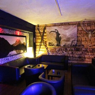 © Heiko Möller m&m bar Ein gemütlicher Innenraum mit dunklen Ledermöbeln und einer Holzverkleidung. An der Wand hängt ein Bild eines Bergpanoramas und ein stilisiertes Ziegenmotiv. | © Heiko Möller m&m bar