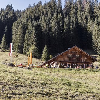 Eine gemütliche Berghütte umgeben von dichten, grünen Wäldern. Im Vordergrund sieht man eine Wiese mit bunten Fähnchen. | © Kleinwalsertal Tourismus | Bastian Morell