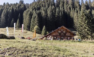 Eine gemütliche Berghütte umgeben von dichten, grünen Wäldern. Im Vordergrund sieht man eine Wiese mit bunten Fähnchen. | © Kleinwalsertal Tourismus | Bastian Morell