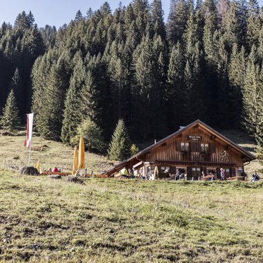 Eine gemütliche Berghütte umgeben von dichten, grünen Wäldern. Im Vordergrund sieht man eine Wiese mit bunten Fähnchen. | © Kleinwalsertal Tourismus | Bastian Morell