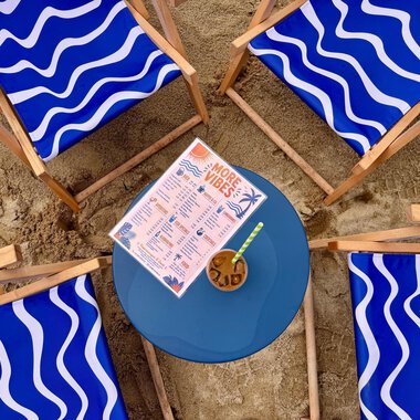 Vier blaue Liegestühle mit Wellendesign stehen im Sand um einen runden Tisch. Auf dem Tisch liegt eine Getränkekarte und ein Cocktail. | © MORE VIBES by Alex Lambsdorff