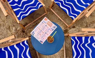 Vier blaue Liegestühle mit Wellendesign stehen im Sand um einen runden Tisch. Auf dem Tisch liegt eine Getränkekarte und ein Cocktail. | © MORE VIBES by Alex Lambsdorff