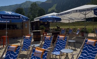 Ein Strandbereich mit blauen Liegestühlen und Sonnenschirmen. Im Hintergrund sind grüne Berge zu sehen. | © MORE VIBES by Alex Lambsdorff