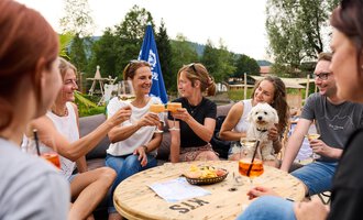 Eine Gruppe von Frauen und Männern sitzt zusammen an einem Tisch und hebt Gläser zum Anstoßen. Ein kleiner Hund sitzt dabei auf dem Tisch. | © Kleinwalsertal Tourismus | Oliver Farys