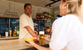 Ein fröhlicher Barkeeper lächelt seinen Kunden am Tresen zu. Im Vordergrund wird eine Portion Pommes mit einem Getränk serviert. | © Kleinwalsertal Tourismus | Oliver Farys