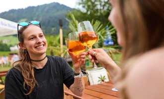 Zwei Frauen stoßen mit Getränken an und lächeln dabei. Im Hintergrund sind grüne Berge und Pflanzen zu sehen. | © Kleinwalsertal Tourismus | Oliver Farys