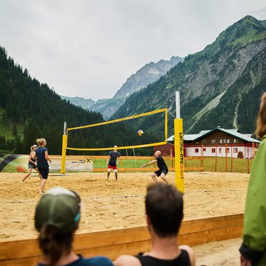 Ein Beachvolleyballspiel in den Bergen, umgeben von einer malerischen Landschaft. Die Spieler sind aktiv auf dem Sandplatz, während Zuschauer zuschauen. | © Kleinwalsertal Tourismus | Oliver Farys