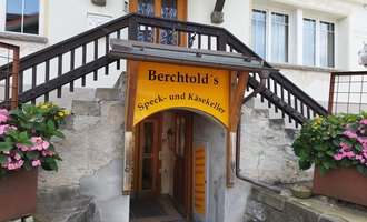An entrance area with colorful signage for Berchtold's Bacon and Cheese Cellar. Stairs lead into the store, surrounded by potted flowers. | © Kleinwalsertal Tourismus | N. Lughammer