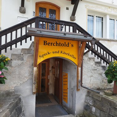 Ein Eingangsbereich zu Berchtold's Speck- und Käsekeller. Über der Tür hängt ein gelbes Schild, umgeben von Blumen. | © Kleinwalsertal Tourismus | N. Lughammer