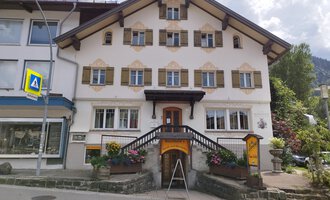 Ein traditionelles alpines Gebäude mit vielen Fenstern und einem geschwungenen Eingang. Bunte Blumen schmücken die Treppe vor dem Haus. | © Kleinwalsertal Tourismus | N. Lughammer