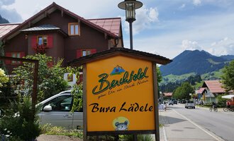 Ein einladendes Schild des "Bura Lädele" in einer malerischen Landschaft. Im Hintergrund sind Berge und ein klarer Himmel zu sehen. | © Kleinwalsertal Tourismus | N. Lughammer