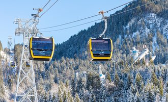Eine Seilbahn mit gelben Gondeln schwebt über schneebedeckte Berge und Wälder. Der Himmel ist klar und blau. | © OBERSTDORF · KLEINWALSERTAL BERGBAHNEN