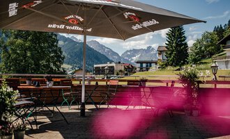Eine gemütliche Terrasse mit einem großen Sonnenschirm und Blick auf die Berge. Grüne Bäume und eine einladende Umgebung schaffen eine entspannte Atmosphäre.