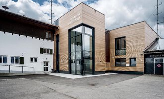 A modern building with wooden elements and large windows. It is located in a rural setting under a cloudy sky. | © Bergrettung Riezlern | Gemeinde Mittelberg