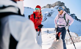 Zwei Frauen stehen im Schnee in den Bergen und lachen. Sie tragen Skikleidung und halten Wanderstöcke. | © Kleinwalsertal Tourismus | Oliver Farys