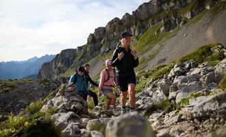 Eine Gruppe von Wanderern erklimmt einen steinigen Pfad in den Bergen. Die Landschaft ist grün und geprägt von beeindruckenden Felsformationen. | © Bergschule Kleinwalsertal @Bergschulel Oliver Farys