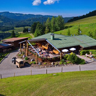 Eine malerische Almhütte umgeben von grünen Wiesen und Wald. Auf der Terrasse genießen Menschen die sonnige Aussicht in die Berge. | © Bergstüble | Jürgen Waffenschmidt