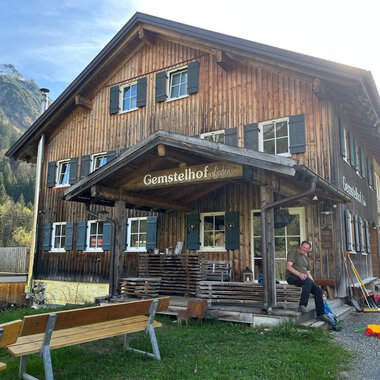 Ein rustikales Holzhaus mit dem Namen "Gemstelhof" und einer einladenden Veranda. Im Vordergrund sieht man eine Person, die an einer Bank sitzt, umgeben von grüner Landschaft und Bergen. | © Bernhards Gemstel Hofladen | Bernhard Heim