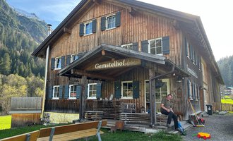 Ein rustikales Holzhaus mit dem Namen "Gemstelhof" und einer einladenden Veranda. Im Vordergrund sieht man eine Person, die an einer Bank sitzt, umgeben von grüner Landschaft und Bergen. | © Bernhards Gemstel Hofladen | Bernhard Heim