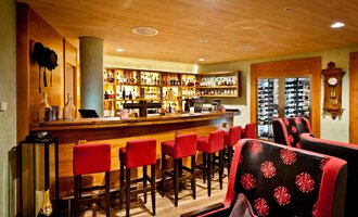 Eine gemütliche Bar mit einem langen Holztheken und roten Barstühlen. Im Hintergrund sind Regale mit Getränken und eine moderne Uhr sichtbar. | © Hotel Birkenhöhe | Fotograf Krug