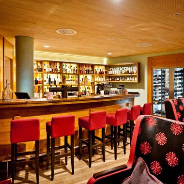 Eine gemütliche Bar mit einem langen Holztheken und roten Barstühlen. Im Hintergrund sind Regale mit Getränken und eine moderne Uhr sichtbar. | © Hotel Birkenhöhe | Fotograf Krug