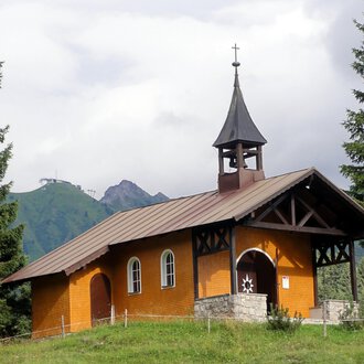 Eine kleine, orangefarbene Kirche steht umgeben von grünen Bäumen. Im Hintergrund sind Berge und eine bewölkte Himmel zu sehen. | © Bruder Klaus Kapelle | Monika Gmeiner