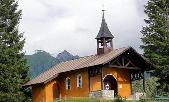 Eine kleine, orangefarbene Kirche steht umgeben von grünen Bäumen. Im Hintergrund sind Berge und eine bewölkte Himmel zu sehen. | © Bruder Klaus Kapelle | Monika Gmeiner