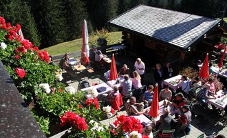 Ein gemütliches Gasthaus in den Bergen mit vielen Gästen im Freien. Rote Blumen und Sonnenschirme verleihen der Umgebung eine einladende Atmosphäre. | © Bühlalpe | Frank Drechsel