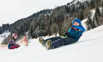 Ein Schlitten liegt im Schnee, umgeben von einer winterlichen Berglandschaft. Die Sonne strahlt hinter den Bergen hervor. | © Kleinwalsertal Tourismus | Oliver Farys