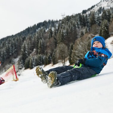 Ein Schlitten liegt im Schnee, umgeben von einer winterlichen Berglandschaft. Die Sonne strahlt hinter den Bergen hervor. | © Kleinwalsertal Tourismus | Oliver Farys