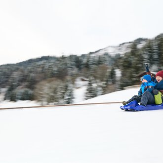 A sled lies in the snow, surrounded by mountains. The sun shines in the background, creating a wintry atmosphere. | © Kleinwalsertal Tourismus | Oliver Farys