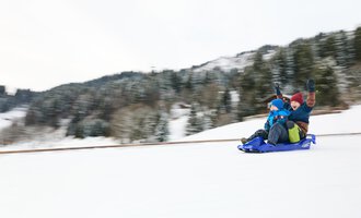 A sled lies in the snow, surrounded by mountains. The sun shines in the background, creating a wintry atmosphere. | © Kleinwalsertal Tourismus | Oliver Farys