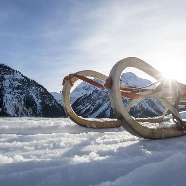 Ein Schlitten liegt im Schnee, umgeben von Bergen. Die Sonne strahlt im Hintergrund und schafft eine winterliche Atmosphäre. | © Kleinwalsertal Tourismus | Dominik Berchtold
