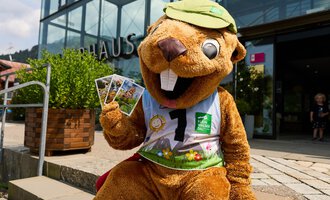 Ein lustiger Wasservogel-Maskottchen sitzt vor einem Gebäude und hält einige Postkarten in der Hand. Es trägt ein grünes Cap und ein buntes Outfit. | © Kleinwalsertal Tourismus | Oliver Farys