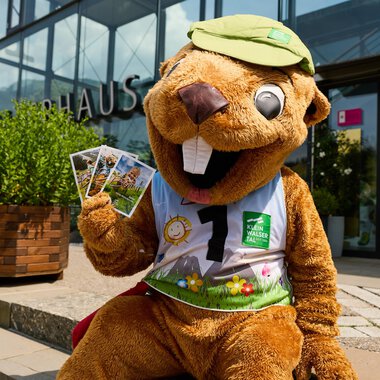 A funny water bird mascot sits in front of a building and holds some postcards in its hand. It is wearing a green cap and a colorful outfit. | © Kleinwalsertal Tourismus | Oliver Farys
