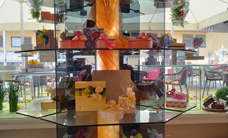 Eine elegante Vitrine mit verschiedenen kunstvoll verzierten Kuchen und Leckereien. Blumen und dekorative Elemente verleihen dem Gesamtbild eine ansprechende Atmosphäre. | © Kleinwalsertal Tourismus | N. Lughammer